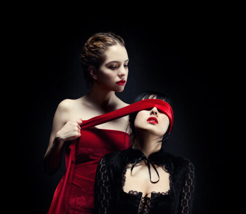 Woman in red dress tying red blindfold on woman in black lace corset